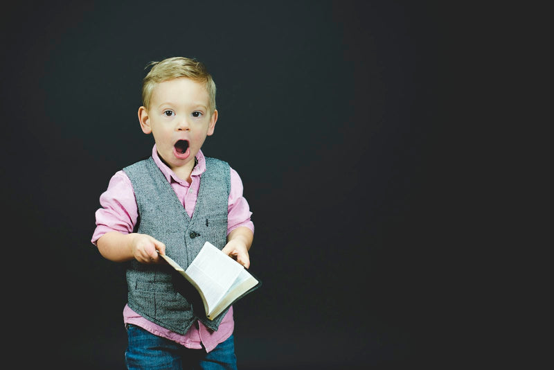 A young boy in a grey vest holding a book, photographed against a dark background.