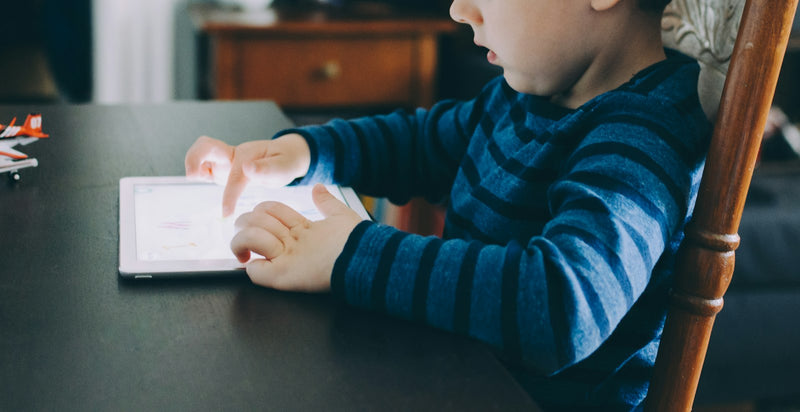 A young child sitting at a table, lit by the glow of a tablet screen in a dim room.