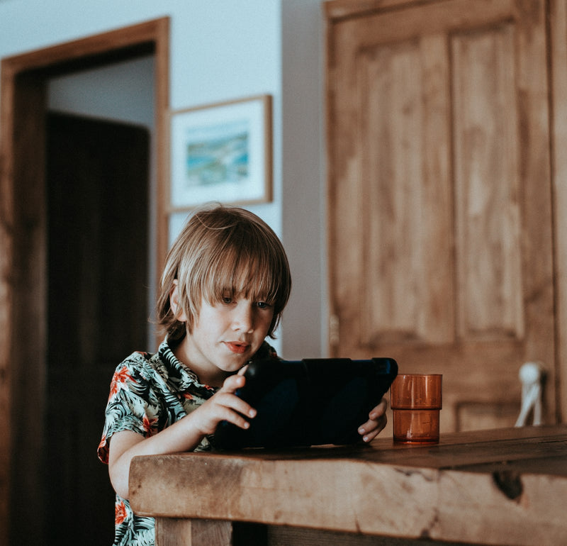 A young child sitting at a wooden table, deeply absorbed in using a tablet, soft natural light on their face.