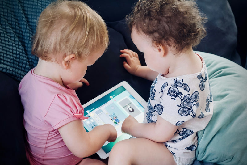 Two toddlers sitting together on a sofa, both absorbed in watching a shared tablet screen
