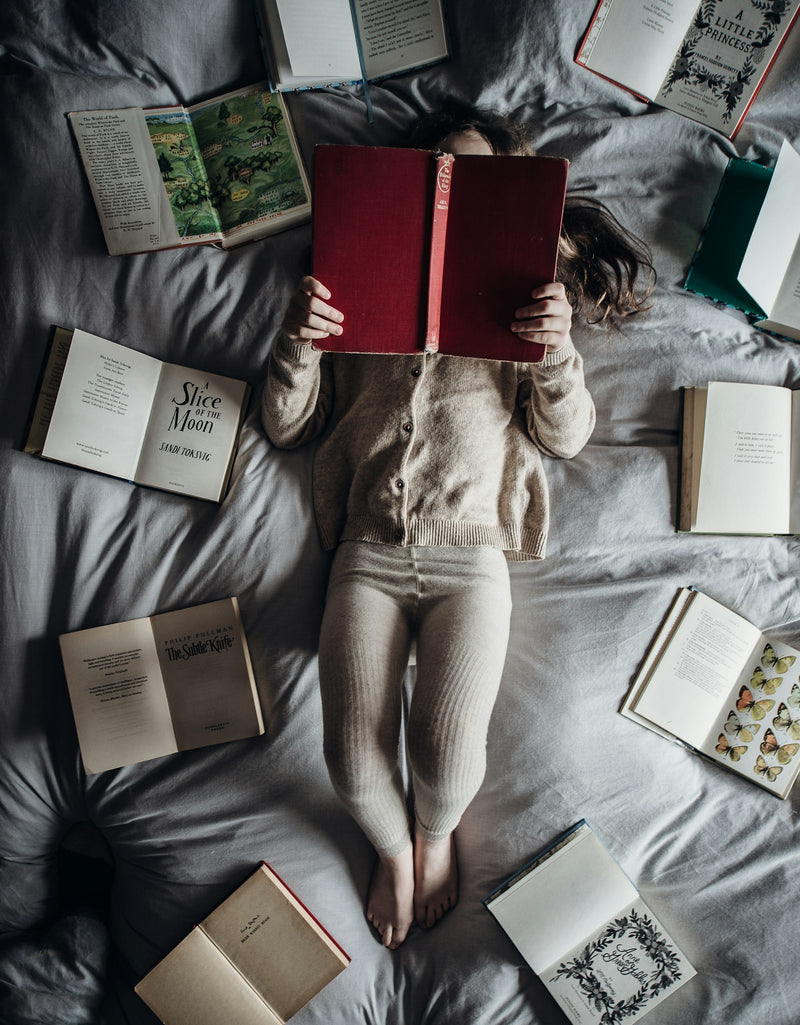 A child lies on a bed surrounded by a pile of books
