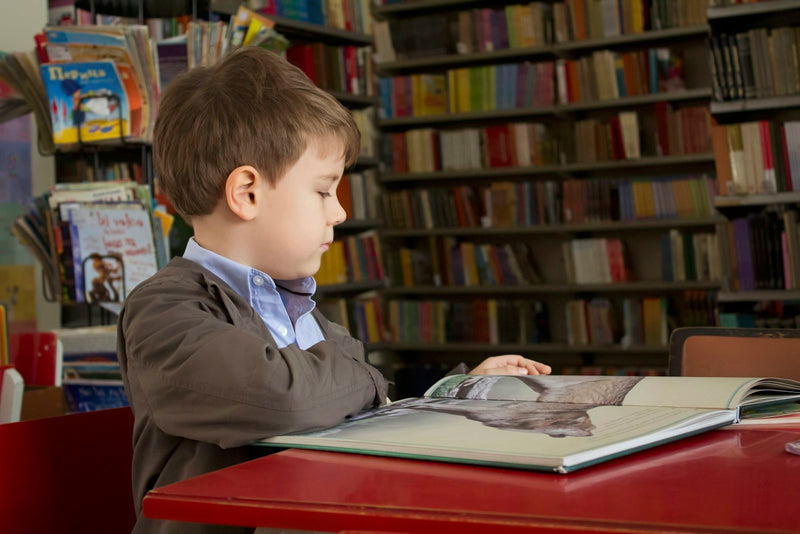 A young boy sitting at a red table, focused on reading a book.