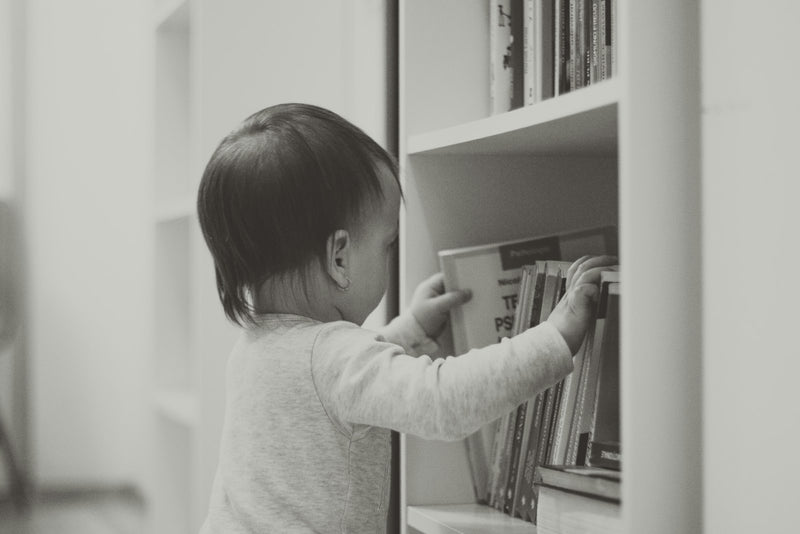 A child standing beside a white wooden bookshelf, looking at the books on it