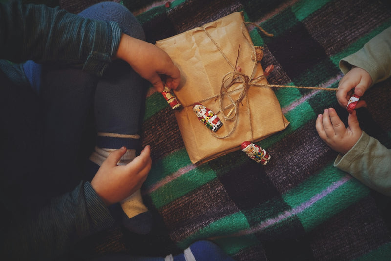 Two young children sit together on the floor, examining a wrapped gift between them.