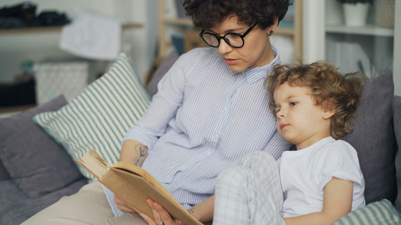 A woman reads a book aloud to a young child while sitting together on a couch.
