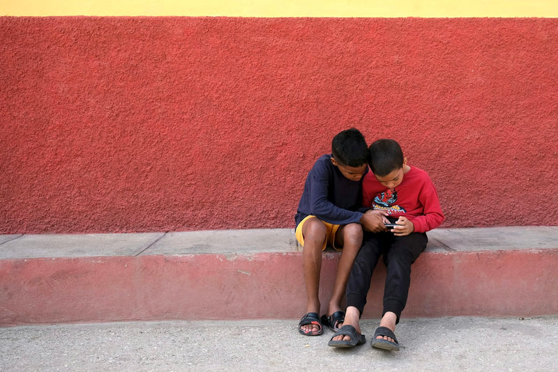 Two boys sitting together outdoors, both looking closely at a phone screen.