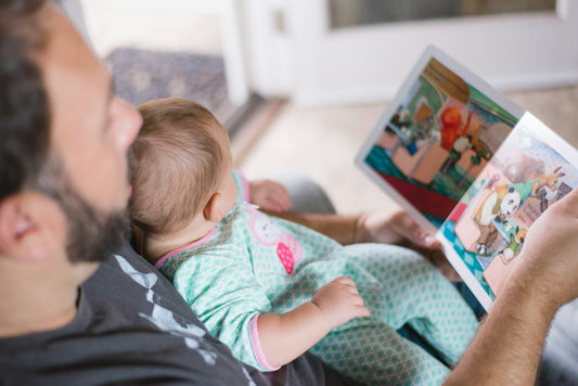 A father sits holding his young child on his lap while reading a storybook together at home.