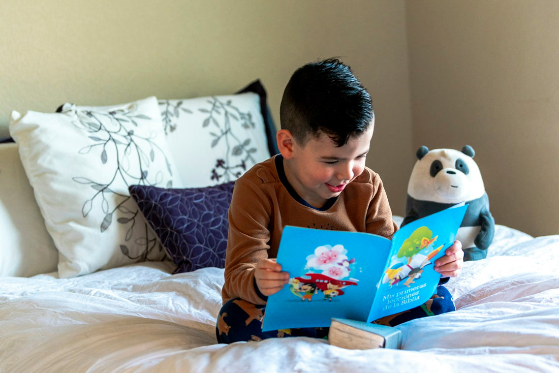 A child lying on a bed reading a book next to a stuffed toy