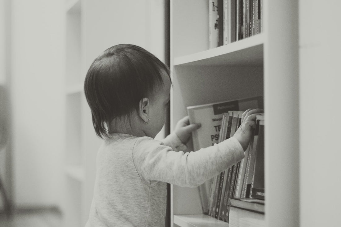 A child standing beside a white wooden bookshelf, looking at the books on it