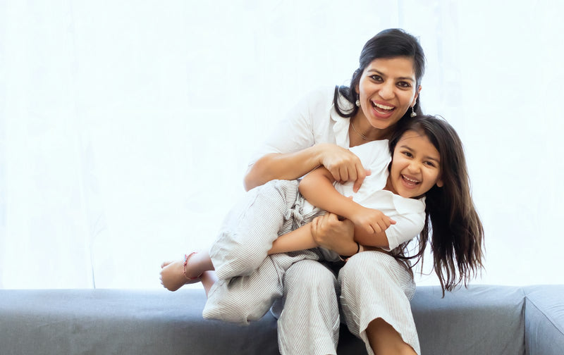 A woman holds a laughing child on a sofa in a bright room, mid-play.