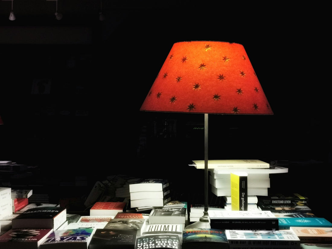 A red table lamp glows beside a stack of books at night, warm light pooling over the covers in an otherwise dark room.