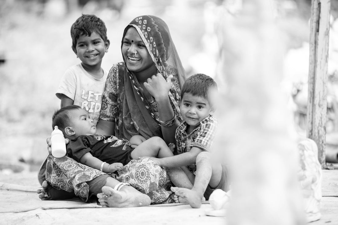 A smiling Indian woman sitting outdoors on the ground with two young children beside her