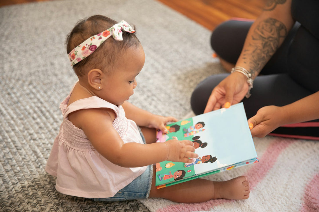 A baby sits on the floor holding open a sturdy board book, exploring its pages.