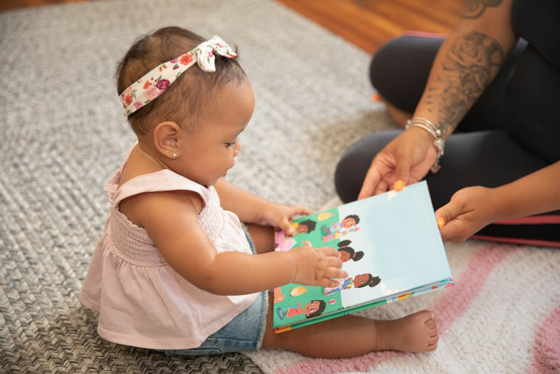 A baby sits on the floor holding open a sturdy board book, exploring its pages.