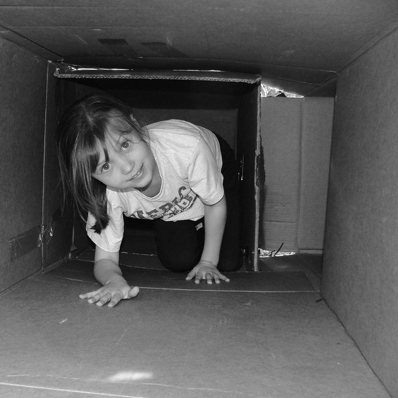 A young child plays inside a cardboard box in a backyard.