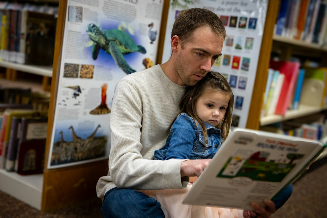A father reads a book aloud to his young daughter in a children's library.