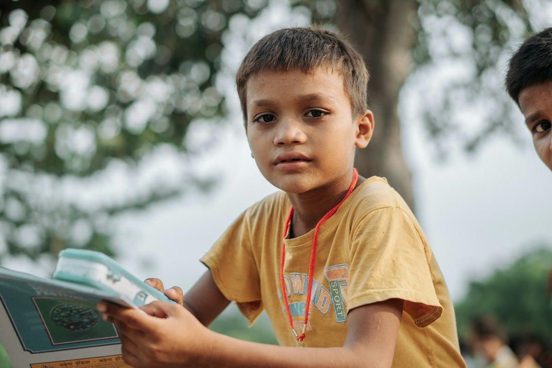 A young boy in a yellow shirt sitting outdoors under a tree, reading a book with other children nearby