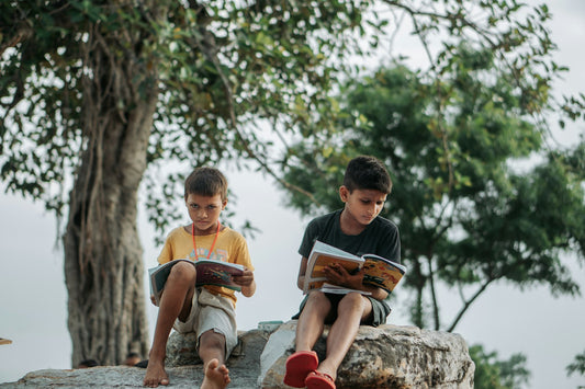 Two young boys sit outdoors under a tree reading books together, fully absorbed in the pages.