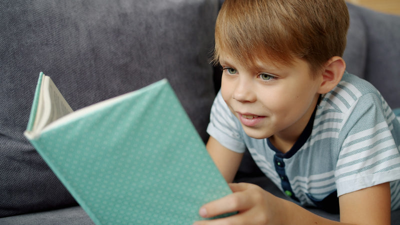 A young boy lying on a couch, absorbed in reading an open book