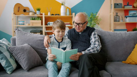 A grandfather and young grandson sitting together on a couch at home, both looking down at an open picture book.