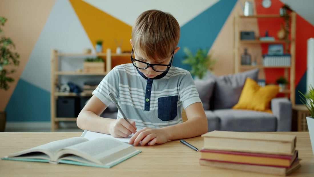A young boy with glasses writing in a notebook at a desk at home.