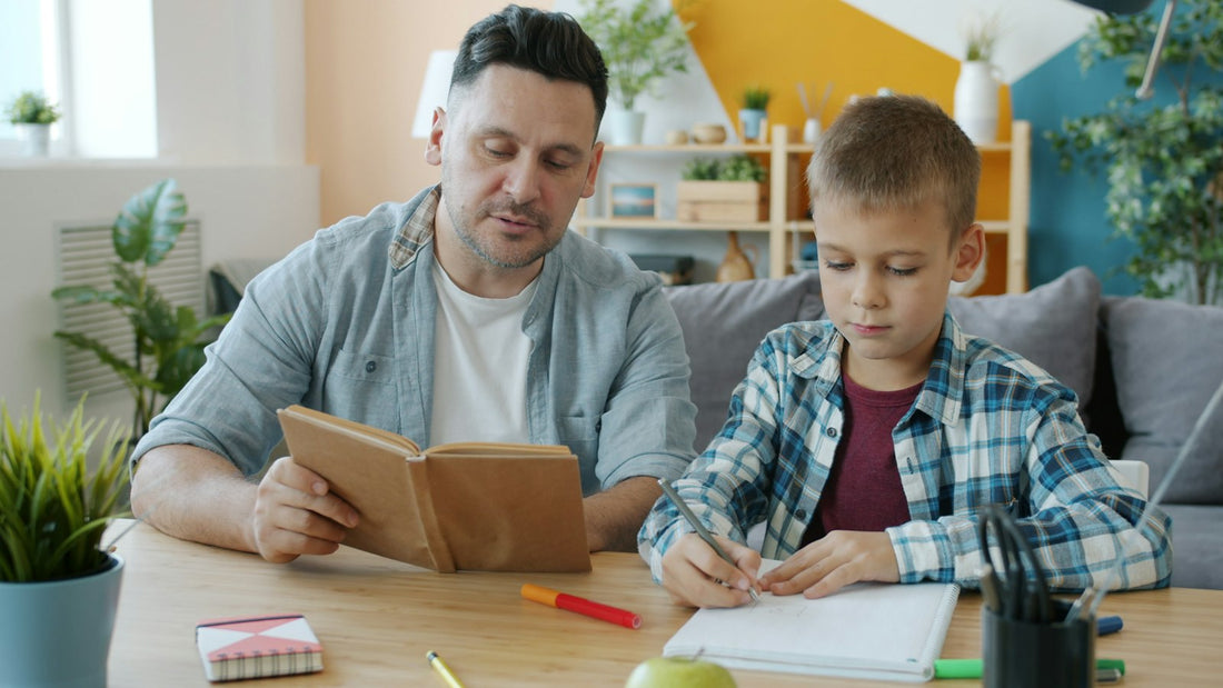 A father reading a book aloud at a table while his young child draws nearby at home