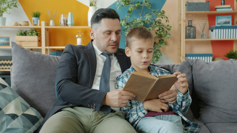 A father reading a book aloud to his young son while they sit together on a couch