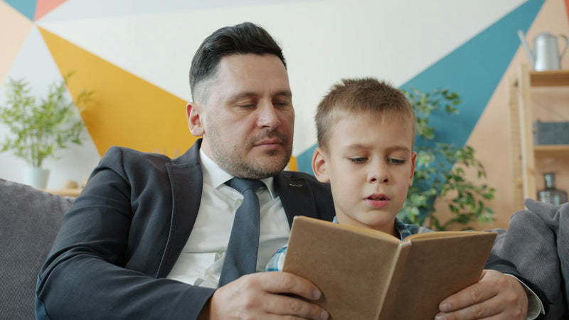A father and young son sitting together on a couch at home, reading a picture book aloud.
