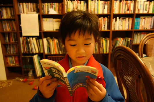 A young child standing in front of tall bookshelves, reading an open book.